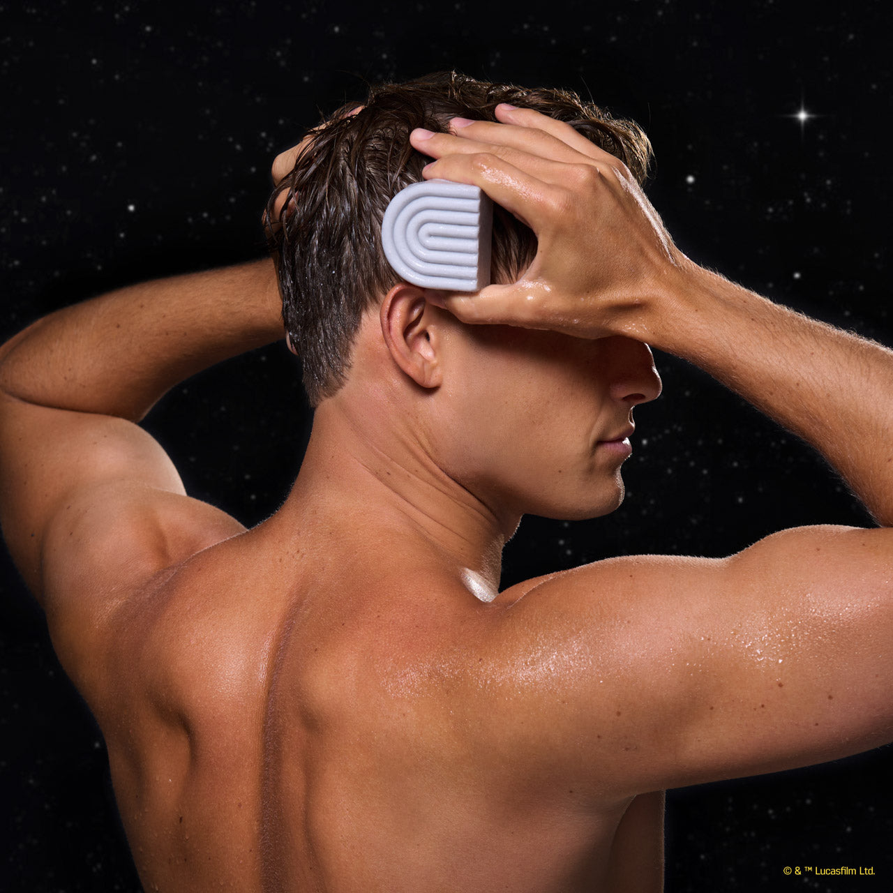 Male model applying KITSCH x Star Wars™ silver-grey S-shaped Rice Water Protein Solid Conditioner Bar (R2-D2 edition) to wet dark hair — side/back lifestyle in-use shot against galaxy starfield backdrop — Strengthening formula For thin, fine, & damaged hair
