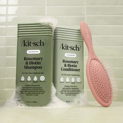 Kitsch Rosemary & Biotin shampoo and conditioner bottles with a pink brush on a tiled bathroom background.
